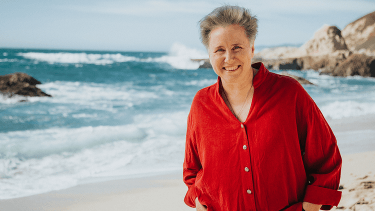 A smiling woman in a red shirt standing on a beach, with the ocean waves and rocky cliffs in the background, creating a serene and natural setting.