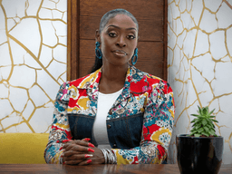 Dilys Sillah seated at a table wearing a colorful floral jacket in a modern interior.