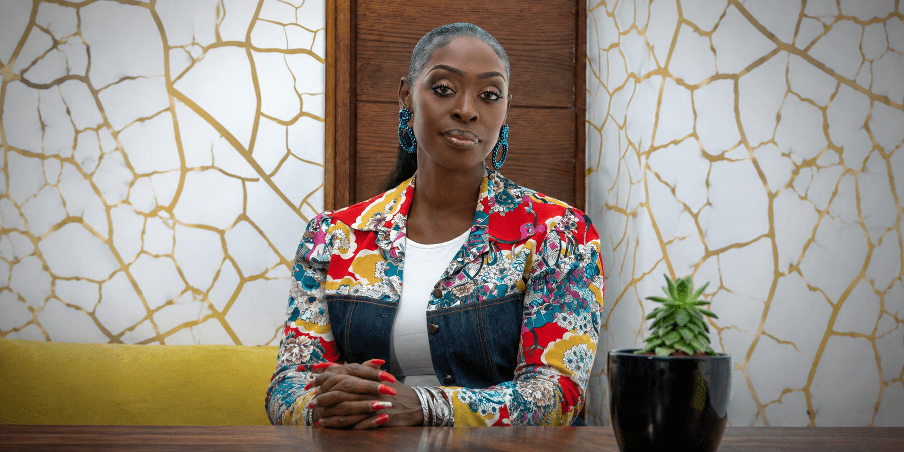 Dilys Sillah seated at a table wearing a colorful floral jacket in a modern interior.