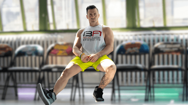 Man in gym attire performing seated L-sit exercise in front of colorful chairs.