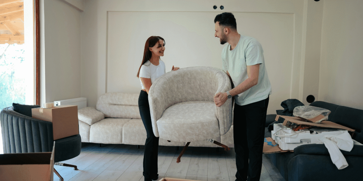 This image shows a man and a woman in the middle of a move or furniture arrangement. They are both smiling and appear to be lifting a modern, beige armchair together. In the background, a spacious living room is being set up, with a couple of couches and cardboard boxes scattered around. The atmosphere feels light-hearted and collaborative, suggesting they are in the process of settling into a new space.