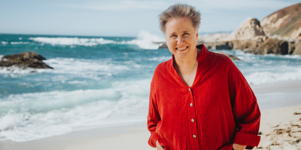 A smiling woman in a red shirt standing on a beach, with the ocean waves and rocky cliffs in the background, creating a serene and natural setting.