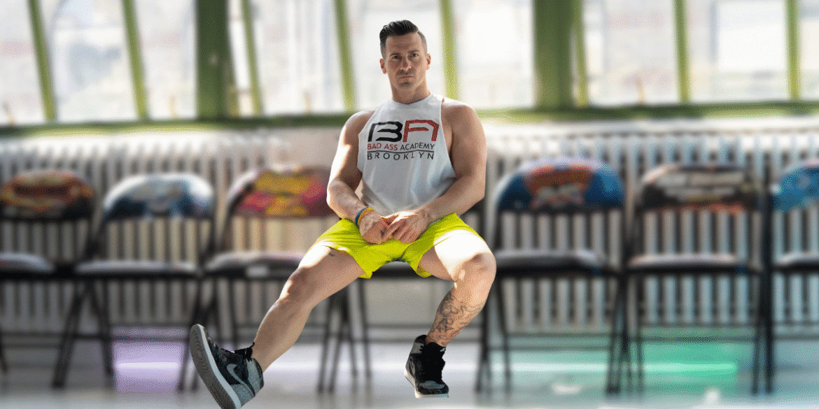 Man in gym attire performing seated L-sit exercise in front of colorful chairs.