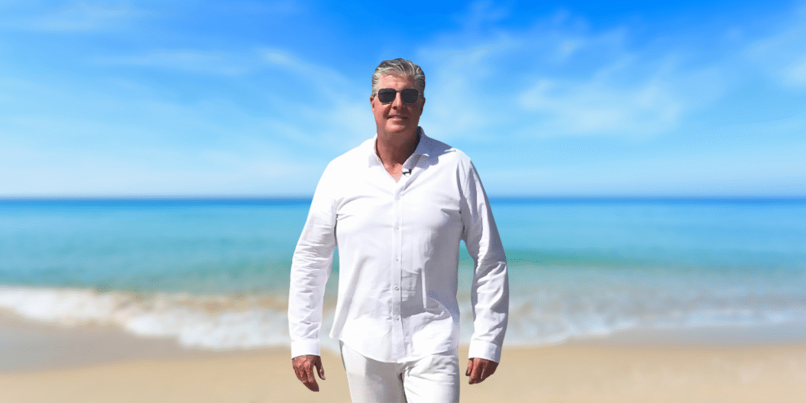 Kevin L. McMullen walks confidently along a beach, dressed in a white shirt and sunglasses, with a serene ocean view in the background.
