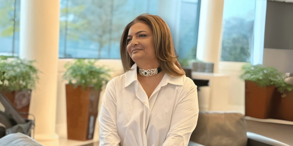 Portrait of a woman with shoulder-length hair wearing a white button-down shirt and a decorative choker necklace, smiling and looking to the side, with potted plants in the background.