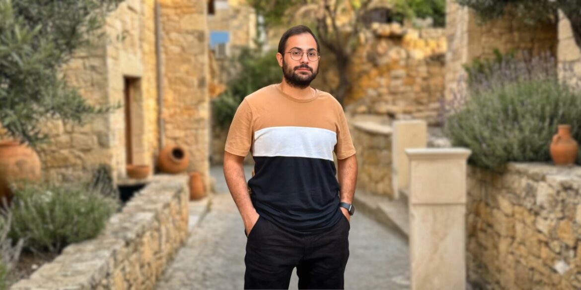 A man with glasses stands in front of a textured stone wall, embodying a thoughtful presence in a poetic setting.
