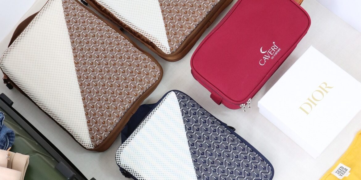 A collection of travel bags on a table, featuring brown and navy patterned cases, a red Caleri pouch, and a white Dior box. The scene conveys organization.