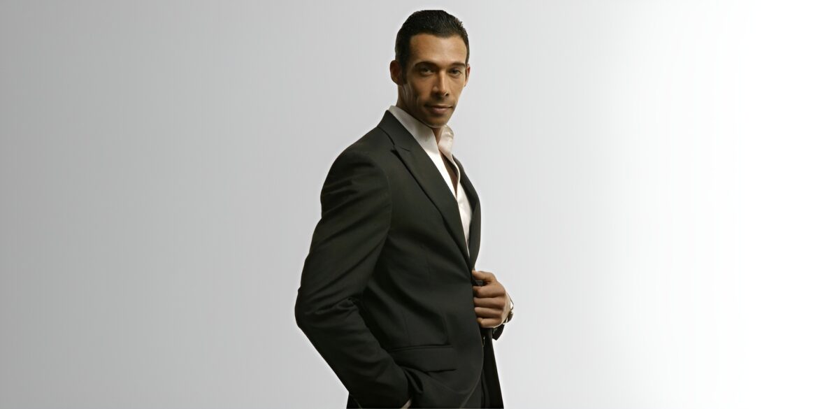 Man in dark suit adjusting jacket, looking confidently at camera against plain background.