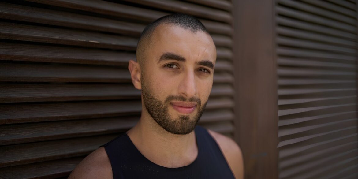 A man with a shaved head and beard gazes calmly at the camera, standing against a textured wooden slat wall. He wears a black tank top, creating a serene tone.
