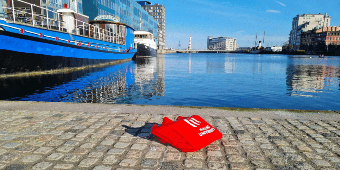 Red Malmö University tote bag displayed beside a waterfront canal in Malmö, Sweden.