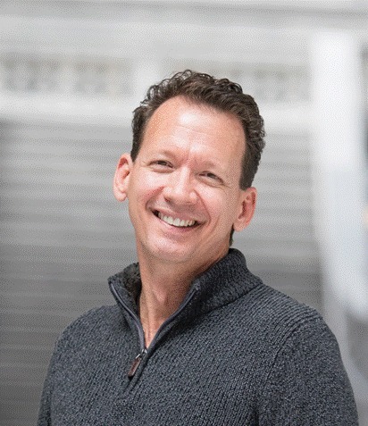 John R. Spender smiling in a gray quarter-zip sweater indoors.