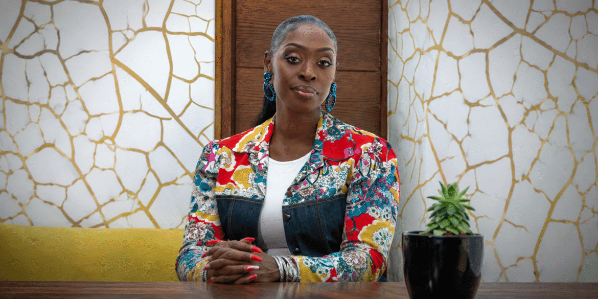Dilys Sillah seated at a table wearing a colorful floral jacket in a modern interior.